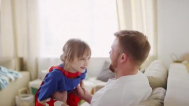 Süper kahraman kostümü giymiş, babasının dizlerinin üstünde koşan, mutlulukla kucaklaşan ve birlikte gülen bir kızın orta boy fotoğrafı.