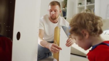 Süper kahraman kostümlü küçük bir çocuğun, babası ve genç kızıyla birlikte beyaz oyuncak roket evi inşa ederken omuz üstünden çekilen fotoğrafı.