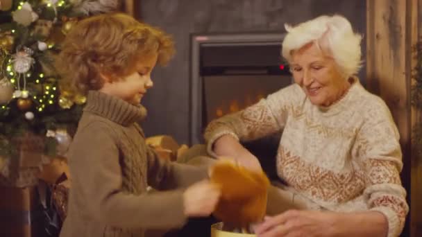 中のショットの笑顔高齢者の女性を助ける小さな男の子Unpackクリスマスギフトボックスと上に置くニットオレンジ冬の帽子とともにザボン笑顔と抱擁 — ストック動画