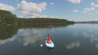 Gölde, bulutları ve ağaçları yansıtan suyla kürek çeken iki kadının yüksek açılı görüntüsü.
