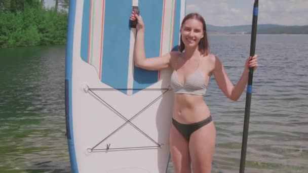 中等比例的迷人微笑的女人 双手拿着桨和苏打水 背对着清澈的湖泊 — 图库视频影像