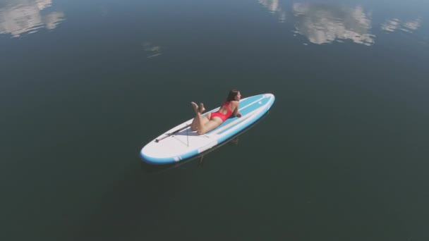 澄んだ青い空の下で開かれた湖で パドルボードの日光浴に横たわっている美しい女性の鳥目の映像 — ストック動画