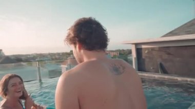 Keyifli bir adam havuzun kenarında oturuyor ve kız arkadaşı güneşli bir günde lüks bir tatil köyünde eğleniyor.