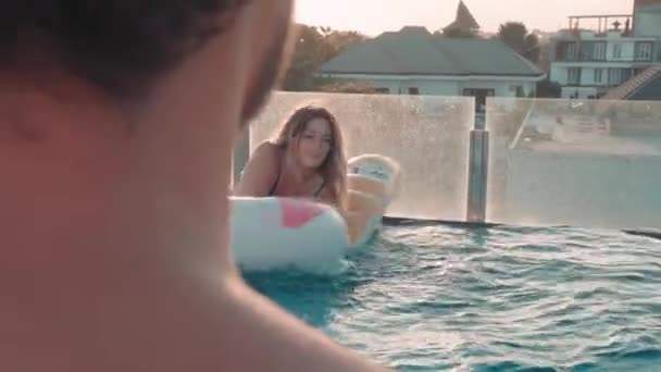 穿着绿松石泳衣的漂亮女孩一边骑着可笑的水床 一边和坐在游泳池边的男朋友聊天 — 图库视频影像