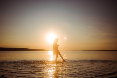 Gün batımının arka planında suda koşan bir kızın silueti. Su her yöne sıçrar. Tasasız bebek eğlencesi. Mutlu sağlıklı bir çocukluk.