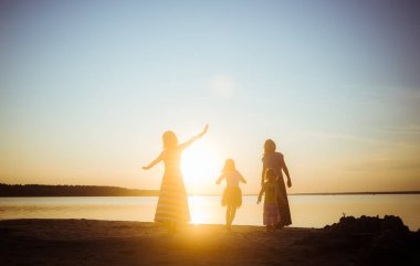 Çocukların siluetleri ve anneleri gün batımında sahilde zıplayıp eğleniyorlar. Genç ve yaşlı nesiller arasında iyi bir ruh hali ve eğlence. Güzel manzara.