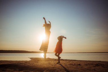 Mutlu anne ve kız gün batımında sahilde zıplayıp dans ediyorlar. İki nesildir iyi ilişkiler içindeyiz. Eğlenceli aile eğlencesi doğada. Oyunlar ve açık hava aktiviteleri yoluyla sağlık tanıtımı.