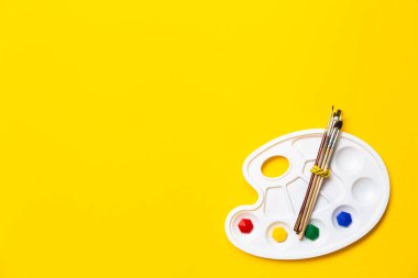 Sarı arka planda ahşap doğal boya fırçaları olan renkli plastik palet. Sanat okulu için metin, hobiler, çizim dersleri, yaratıcılığı ifade etme veya ilham eksikliği.