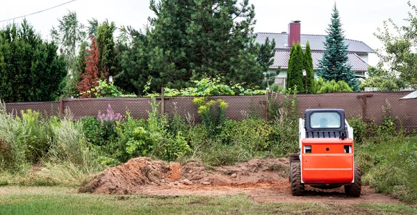 A skid steer loader clears the site for construction. Land work by the territory improvement. Machine for work in confined areas. Small tractor with a bucket for moving soil, turf and bulk materials.