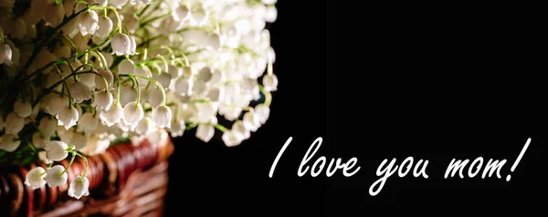 Postcard with the phrase I love you mom. Banner with large beautiful bouquet of white lilies of the valley, packed in a brown wicker basket and isolated on a black background.