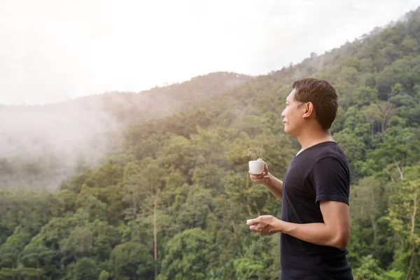 Asia man wearing a black shirt holding a cup of coffee with smoke backdrop mountain nature.