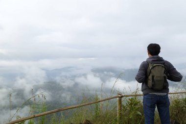 Orta yaşlı Asya adam sırt çantası rahatla ve yenileme dağ zemin üzerine yüksek dağlar, beyaz bulutlar ve sis bir manzara olduğunu.
