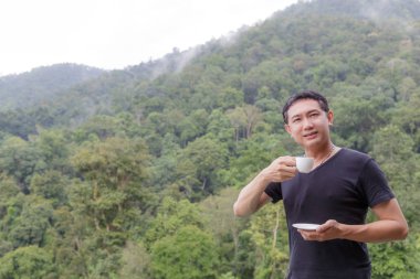 Bir fincan kahve duman zemin dağ doğa ile tutarak siyah tişört giyen Asya adam.