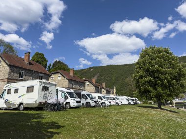 Montherme, France, 05 Mayıs 2025: Fransız kırsalında çimenli bir alana park etmiş beyaz karavan araçları, arka planda eski taş evler ve yeşil tepeler, güneşli bir günün tadını çıkarıyorlar.