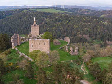 Luc kalesinin ve şapelin görkemli harabeleri bir tepenin üzerinde duruyor, arka planda görünür rüzgar türbinleriyle çevrili yeşil bir manzara.