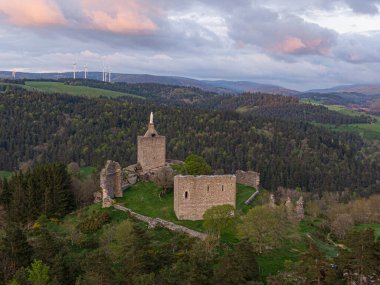 Luc kalesinin kalıntıları günbatımında ormanlık tepelere hükmediyor, rüzgar türbinleri uzaktaki yenilenebilir enerjiyi simgeliyor.