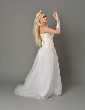 tam uzunlukta portre sarışın kız beyaz fantezi elbise giyiyor. yan profil, gri studio arka plan ayakta poz.