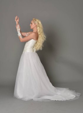 tam uzunlukta portre sarışın kız beyaz fantezi elbise giyiyor. yan profil, gri studio arka plan ayakta poz.