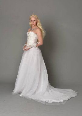 tam uzunlukta portre sarışın kız beyaz fantezi elbise giyiyor. yan profil, gri studio arka plan ayakta poz.