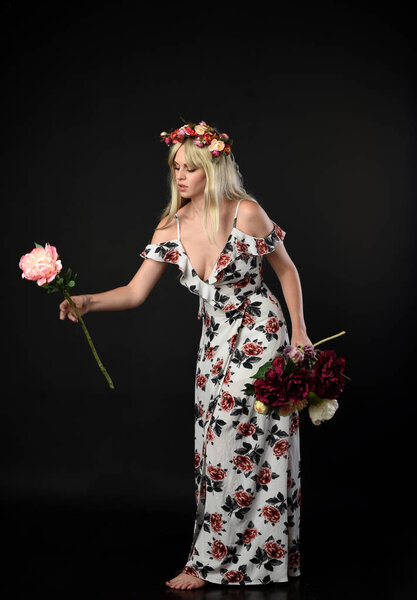 full length portrait of blonde girl wearing floral dress and a flower crown. standing pose against a black studio background.