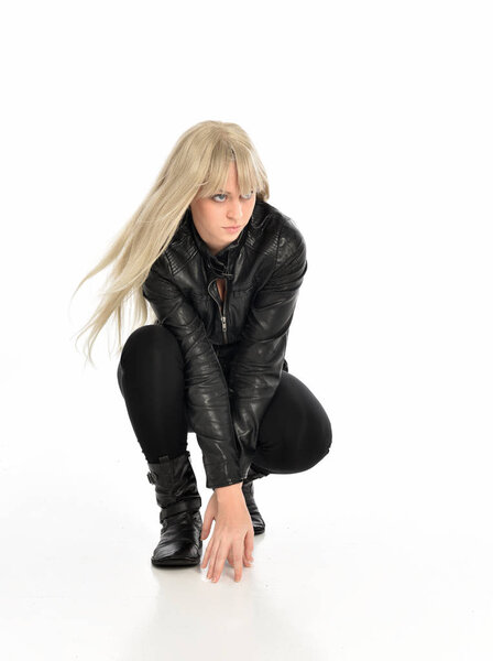 full length portrait of a blonde girl wearing black leather outfit, sitting pose. isolated on white studio background.