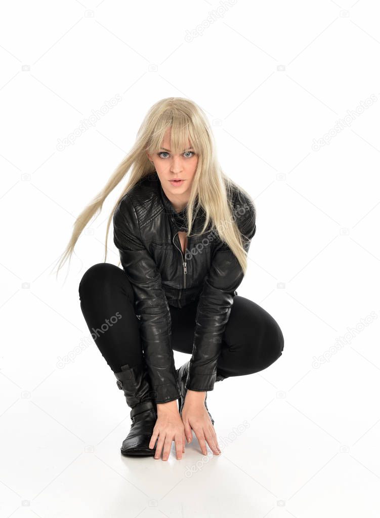 full length portrait of a blonde girl wearing black leather outfit, sitting pose. isolated on white studio background.
