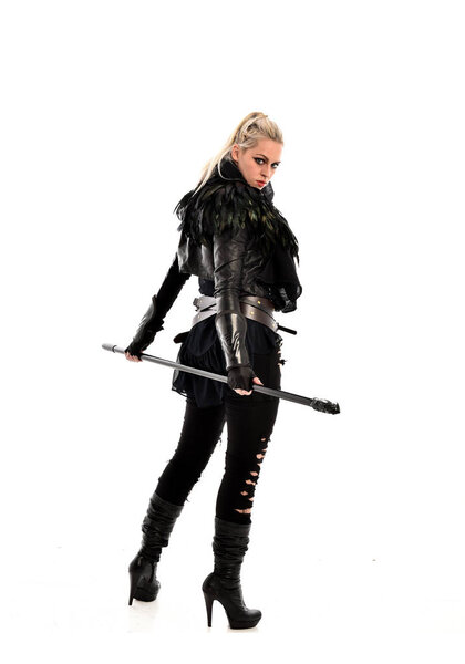 full length portrait of blonde girl wearing black torn outfit, holding a staff. standing pose with back to the camera. isolated on white studio background.