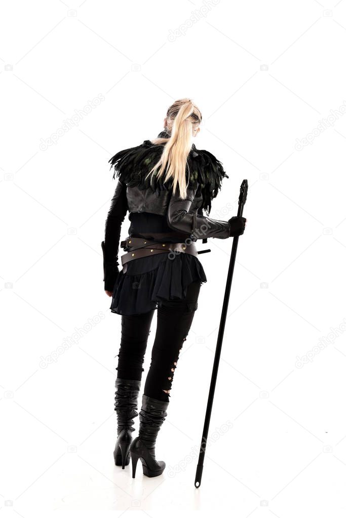 full length portrait of blonde girl wearing black torn outfit, holding a staff. standing pose with back to the camera. isolated on white studio background.