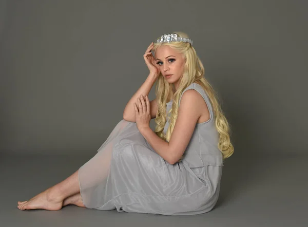 full length portrait of blonde woman wearing crown and pale blue dress. seated pose against a grey studio background.
