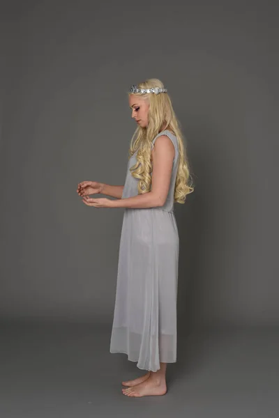 full length portrait of blonde girl wearing crow and grey dress, standing pose in side profile.  grey studio background.