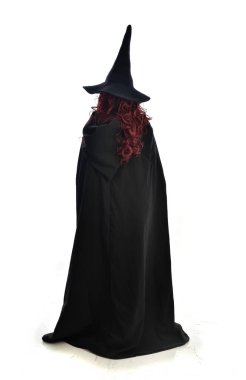 tam uzunlukta uzun siyah pelerin, sivri şapka ve cadı kostümü giyen kırmızı saçlı kız portresi. ayakta poz, beyaz stüdyo arka plan üzerinde izole.