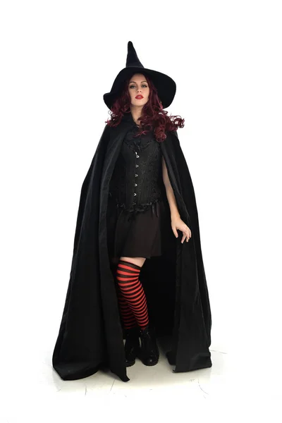 全长肖像的红头发的女孩身穿长黑斗篷 尖帽和巫婆服装 站立的姿态 被隔绝在白色演播室背景 — 图库照片