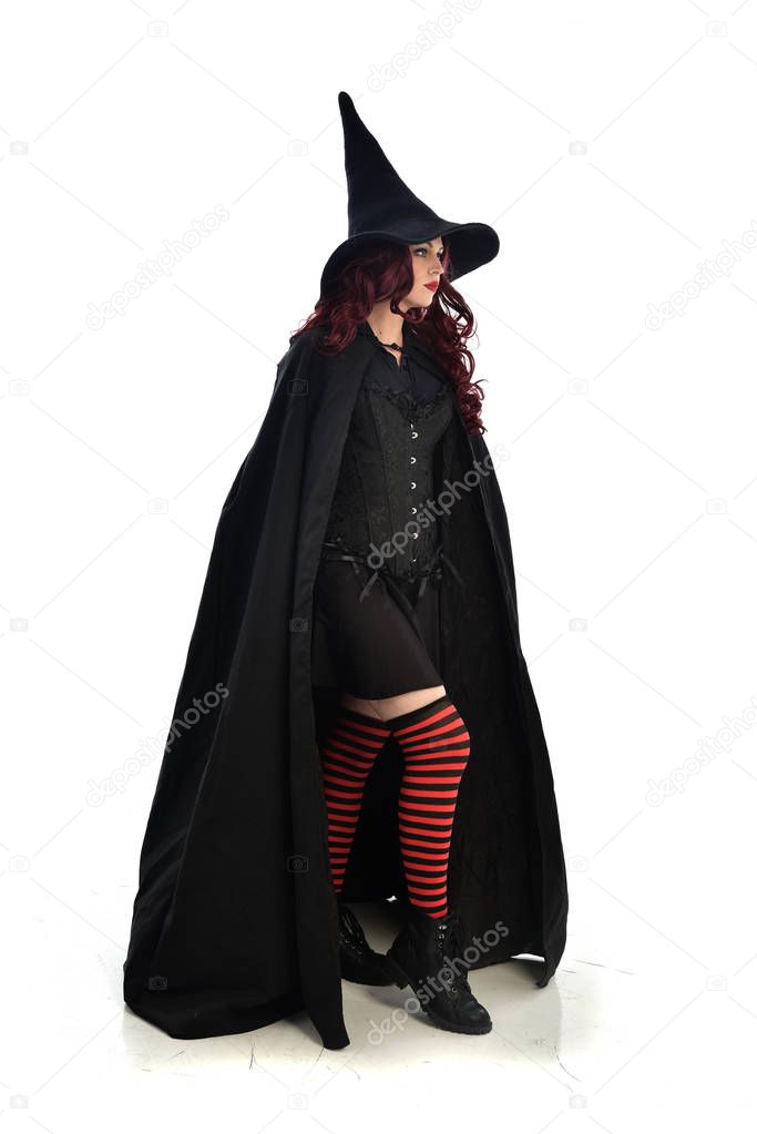 full length portrait of red haired girl wearing long black cloak, pointy hat and witch costume. standing pose, isolated on white studio background.