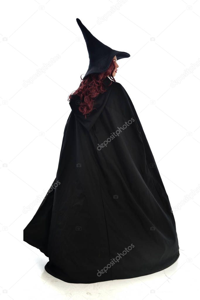 full length portrait of red haired girl wearing long black cloak, pointy hat and witch costume. standing pose, isolated on white studio background.
