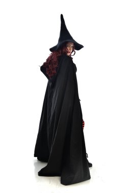 tam uzunlukta uzun siyah pelerin, sivri şapka ve cadı kostümü giyen kırmızı saçlı kız portresi. ayakta poz, beyaz stüdyo arka plan üzerinde izole.