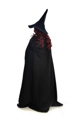 tam uzunlukta uzun siyah pelerin, sivri şapka ve cadı kostümü giyen kırmızı saçlı kız portresi. ayakta poz, beyaz stüdyo arka plan üzerinde izole.