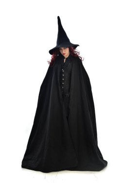 tam uzunlukta uzun siyah pelerin, sivri şapka ve cadı kostümü giyen kırmızı saçlı kız portresi. ayakta poz, beyaz stüdyo arka plan üzerinde izole.