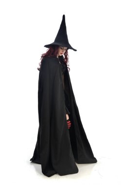 tam uzunlukta uzun siyah pelerin, sivri şapka ve cadı kostümü giyen kırmızı saçlı kız portresi. ayakta poz, beyaz stüdyo arka plan üzerinde izole.