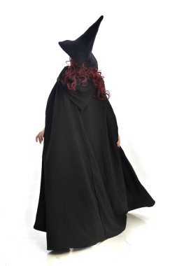 tam uzunlukta uzun siyah pelerin, sivri şapka ve cadı kostümü giyen kırmızı saçlı kız portresi. ayakta poz, beyaz stüdyo arka plan üzerinde izole.