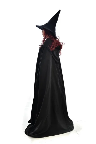 長い黒マント とんがり帽子を被った 魔女の衣装を身に着けている赤髪の女の子の完全な長さの肖像画 立ちポーズ 白いスタジオ背景に分離 — ストック写真