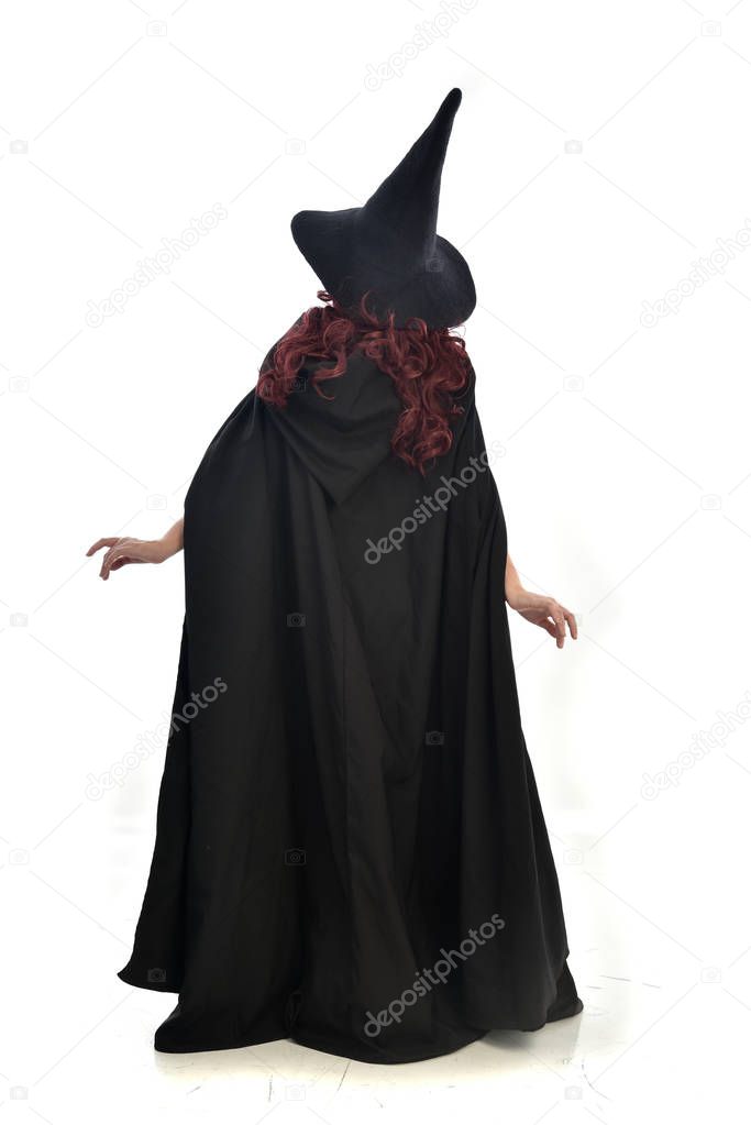 full length portrait of red haired girl wearing long black cloak, pointy hat and witch costume. standing pose, isolated on white studio background.