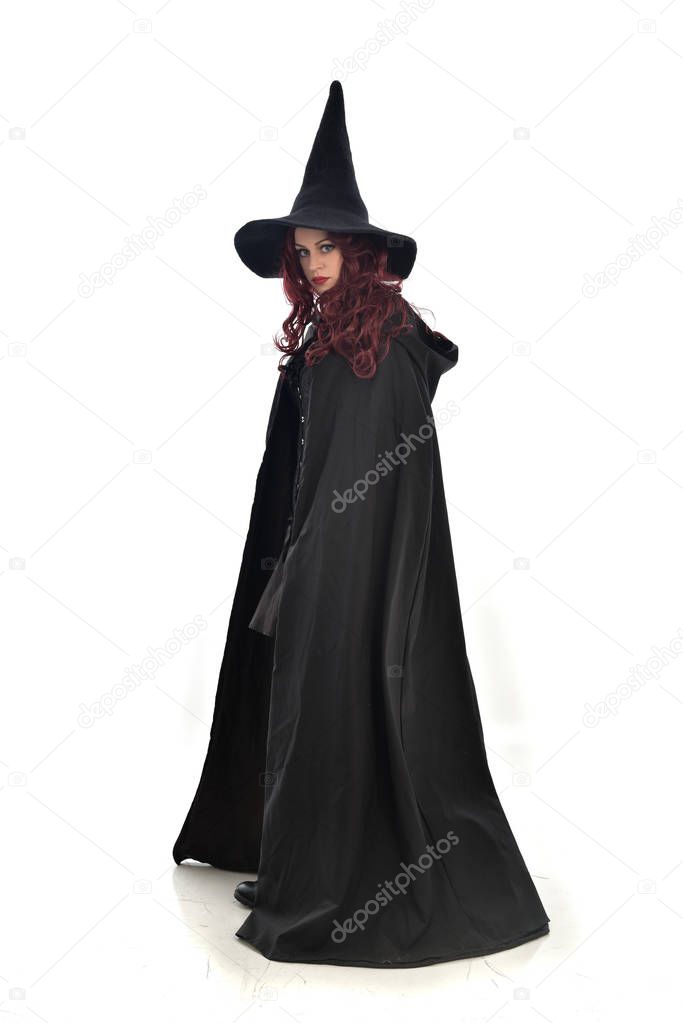 full length portrait of red haired girl wearing long black cloak, pointy hat and witch costume. standing pose, isolated on white studio background.