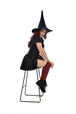 tam uzunlukta uzun siyah pelerin, sivri şapka ve cadı kostümü giyen kırmızı saçlı kız portresi. beyaz stüdyo arka plan üzerinde izole sandalyeye oturmuş poz.