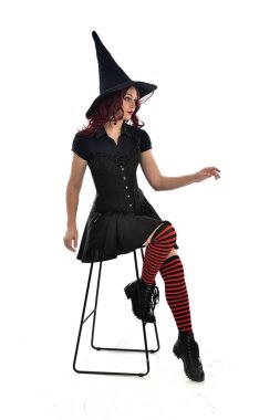 tam uzunlukta uzun siyah pelerin, sivri şapka ve cadı kostümü giyen kırmızı saçlı kız portresi. beyaz stüdyo arka plan üzerinde izole sandalyeye oturmuş poz.