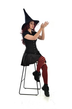 tam uzunlukta uzun siyah pelerin, sivri şapka ve cadı kostümü giyen kırmızı saçlı kız portresi. beyaz stüdyo arka plan üzerinde izole sandalyeye oturmuş poz.