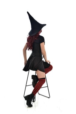 tam uzunlukta uzun siyah pelerin, sivri şapka ve cadı kostümü giyen kırmızı saçlı kız portresi. beyaz stüdyo arka plan üzerinde izole sandalyeye oturmuş poz.