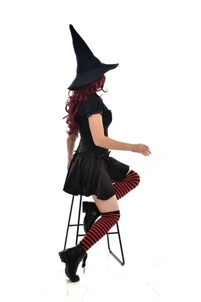 全长肖像的红头发的女孩身穿长黑斗篷 尖帽和巫婆服装 坐在椅子上的姿势 在白色工作室背景下隔离 — 图库照片