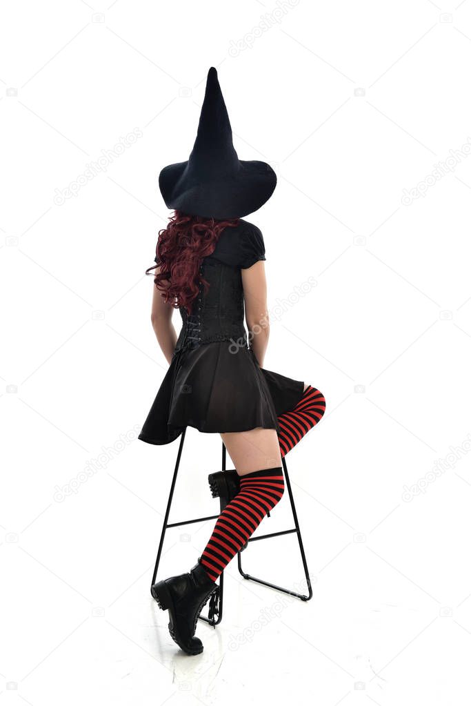 full length portrait of red haired girl wearing long black cloak, pointy hat and witch costume. seated pose on chair, isolated on white studio background.