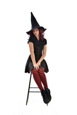tam uzunlukta uzun siyah pelerin, sivri şapka ve cadı kostümü giyen kırmızı saçlı kız portresi. beyaz stüdyo arka plan üzerinde izole sandalyeye oturmuş poz.