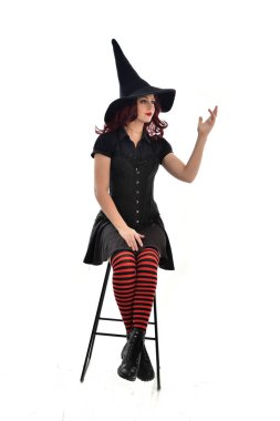 tam uzunlukta uzun siyah pelerin, sivri şapka ve cadı kostümü giyen kırmızı saçlı kız portresi. beyaz stüdyo arka plan üzerinde izole sandalyeye oturmuş poz.